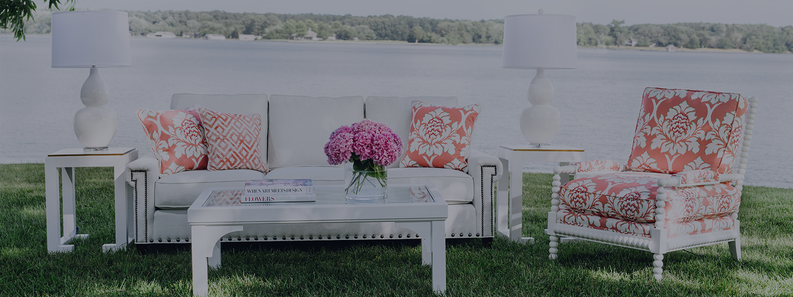 End tables, coffee table and lamps from the Jamie Merida Collection paired with a white sofa and accent chair. On green grass by the water.
